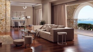 A spacious living area with a large sofa, a coffee table, and a round side table. There is a stone wall and a view of the sea through an arched window.
