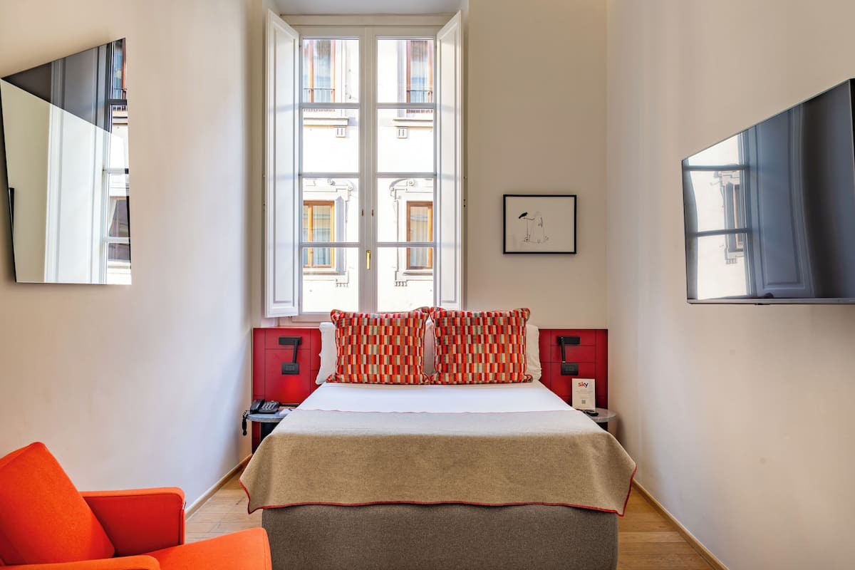 Habitación de hotel con un ventanal grande, cama con almohadones estampados, un sillón rojo, mesita de noche con teléfono y televisión de pantalla plana montada en la pared.