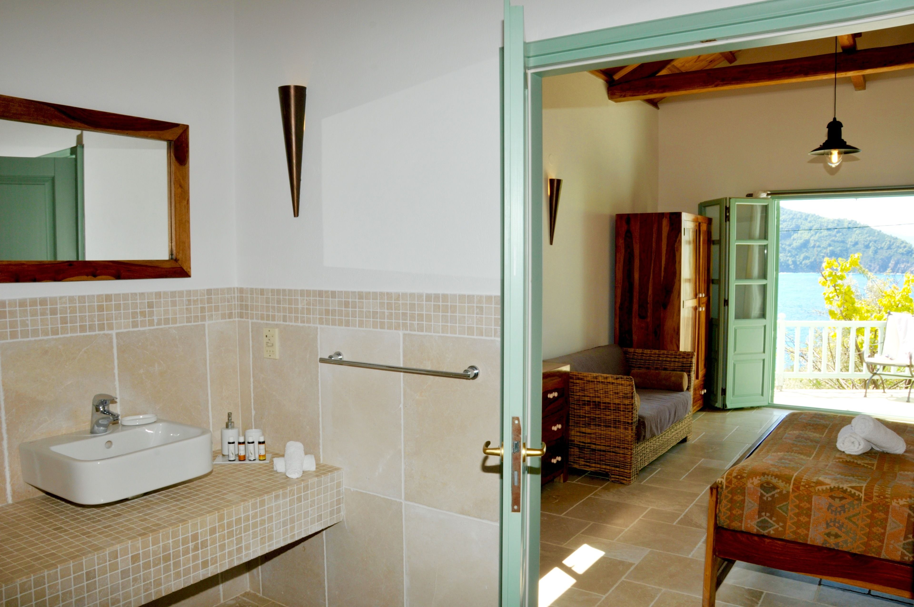 A bathroom with a sink, mirror, and towel rack. There is a door leading to a balcony with a view of the sea.