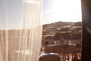 A desert dining area with tables and chairs, a decorative centerpiece, and a view of the desert landscape. - Kam Kam Dunes (Rissani)