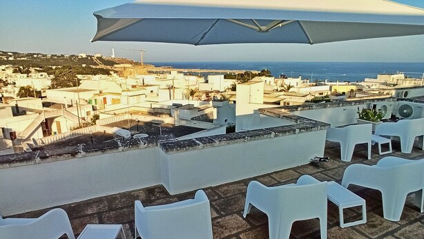 Una terrazza con sedie di plastica bianca e un ombrellone, con vista su una località costiera, edifici e il mare.