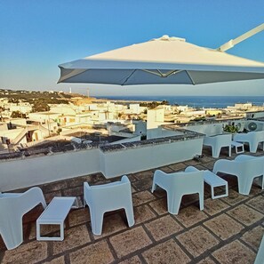 A terrace with white plastic chairs and a large umbrella, overlooking a coastal town with buildings and the sea. - Residence Bellevue (Castrignano del Capo)