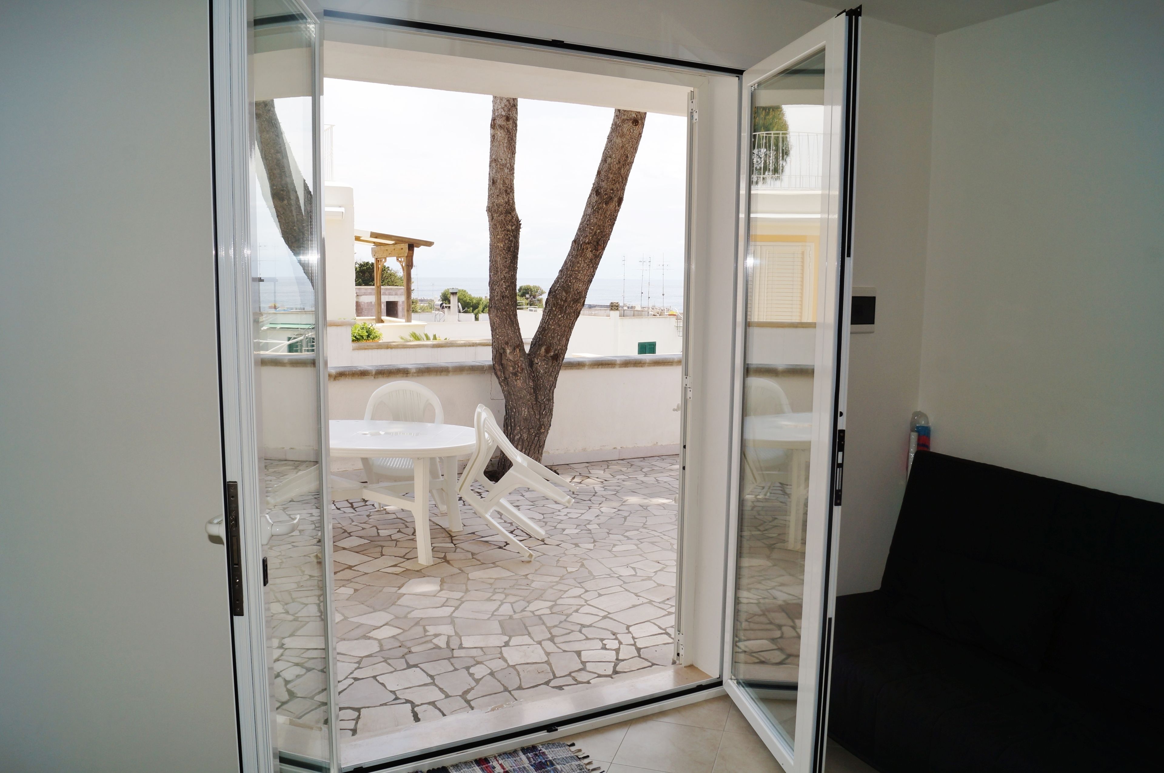 A room with a sliding glass door leading to an outdoor area with a tiled patio, a white table, and two white chairs.