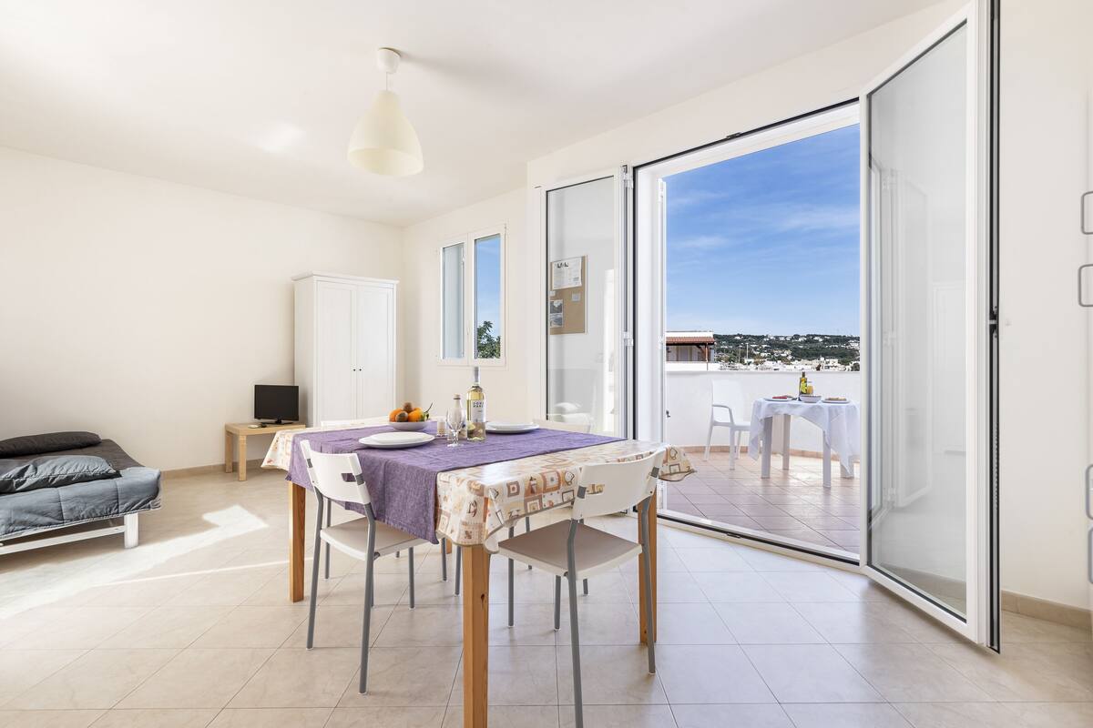 A modern apartment with a dining area, a bed, and a balcony with a city view.