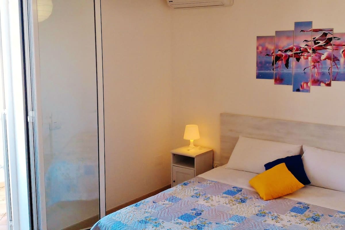 A hotel room with a bed, a nightstand with a lamp, a wall art of flamingos, and a window with a view of the outside.