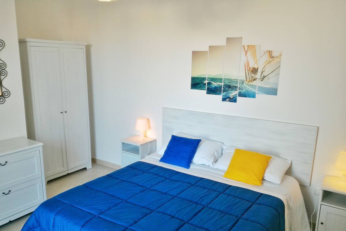 A bedroom with a large bed, a wardrobe, a nightstand with a lamp, and a wall-mounted artwork.
