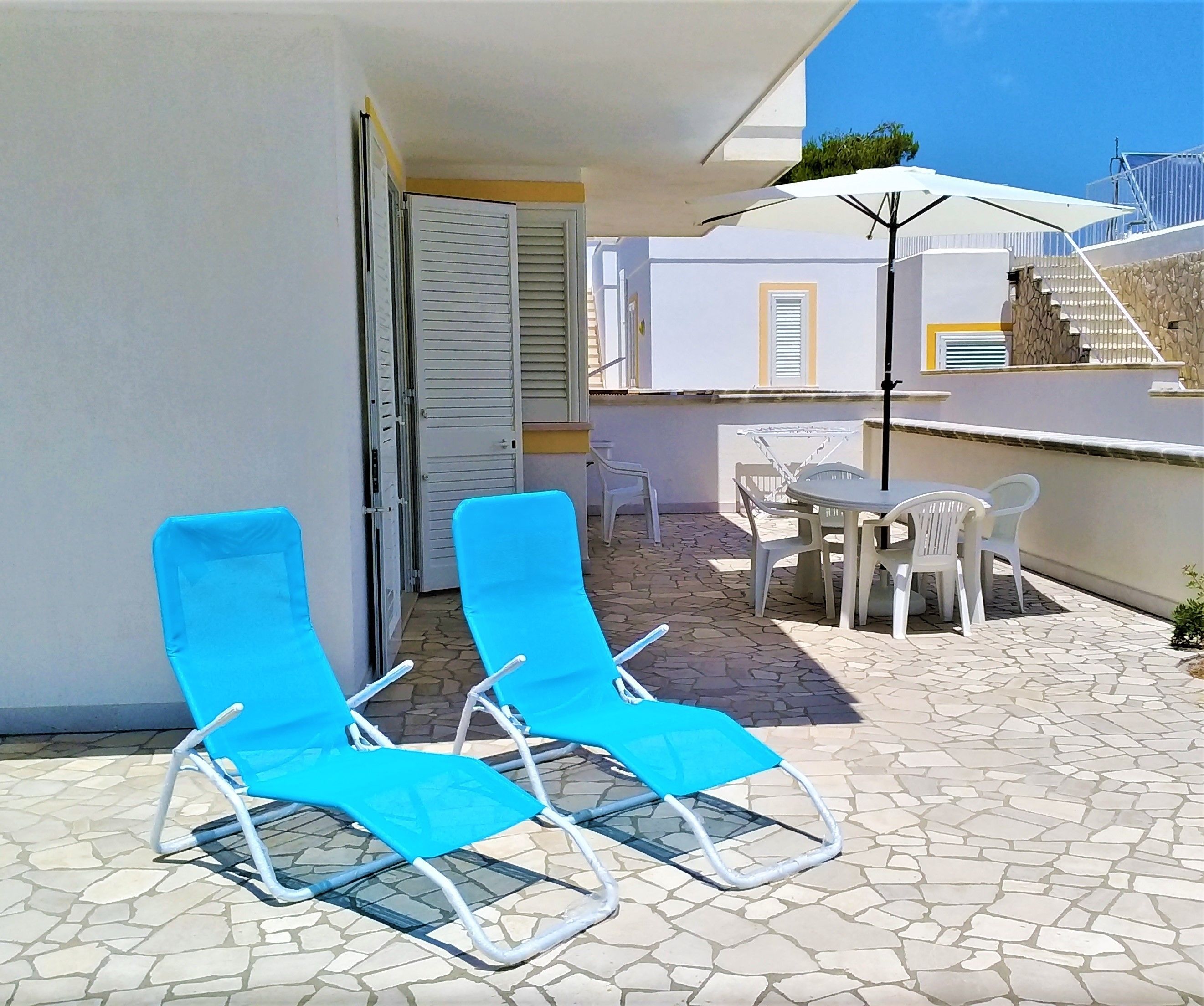 A balcony with two blue lounge chairs, a table with chairs, and a white umbrella.