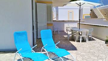 Varanda com duas espreguiçadeiras azuis, uma mesa e cadeiras, e um guarda-sol branco.