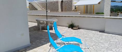 Un patio en la azotea con una mesa y sillas azules, piso de baldosas de piedra y un edificio blanco con un muro de piedra al fondo.
