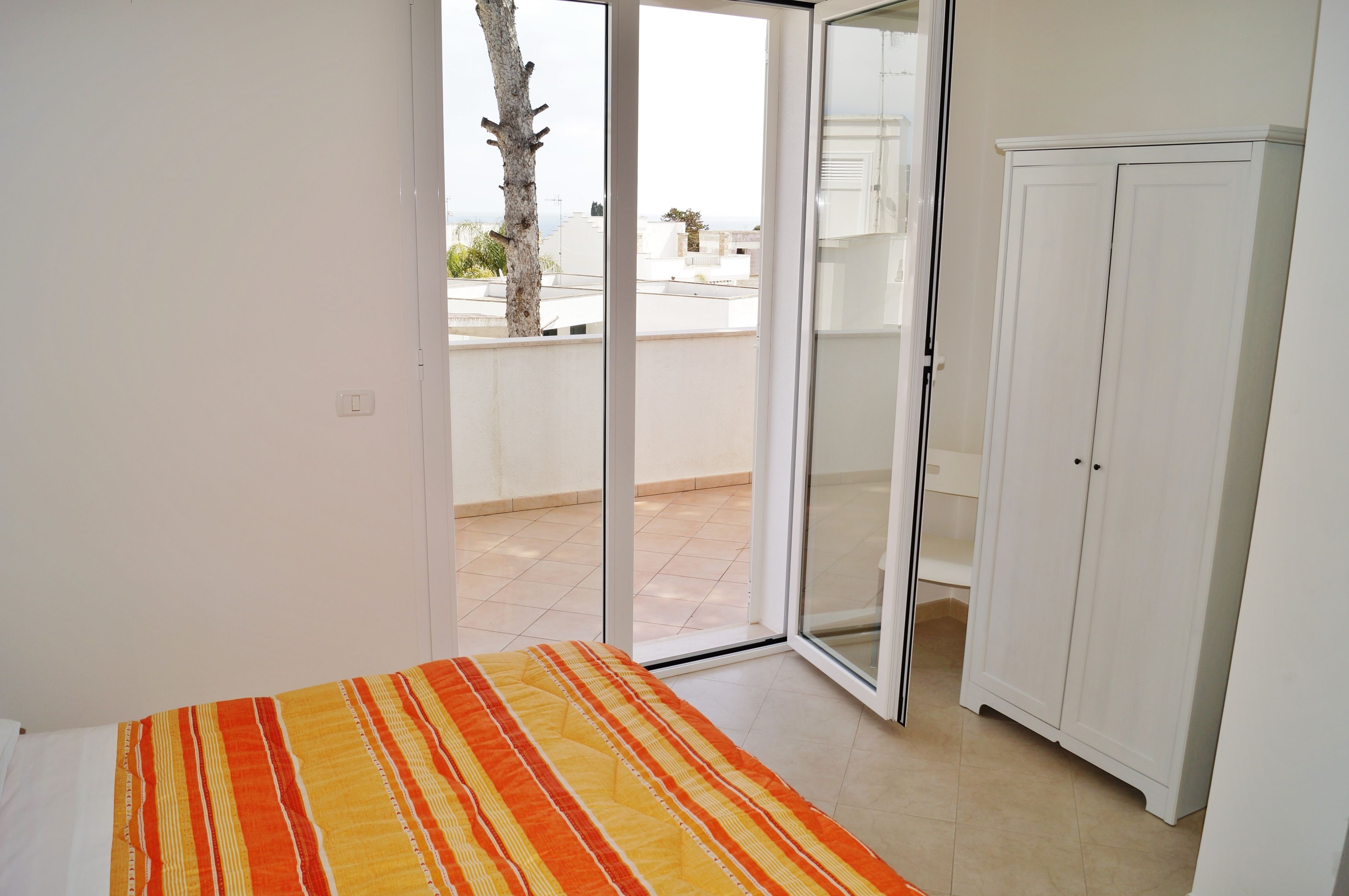 Habitación con cama, armario y puerta corrediza de vidrio que da a un espacio exterior con vistas a edificios y un árbol.