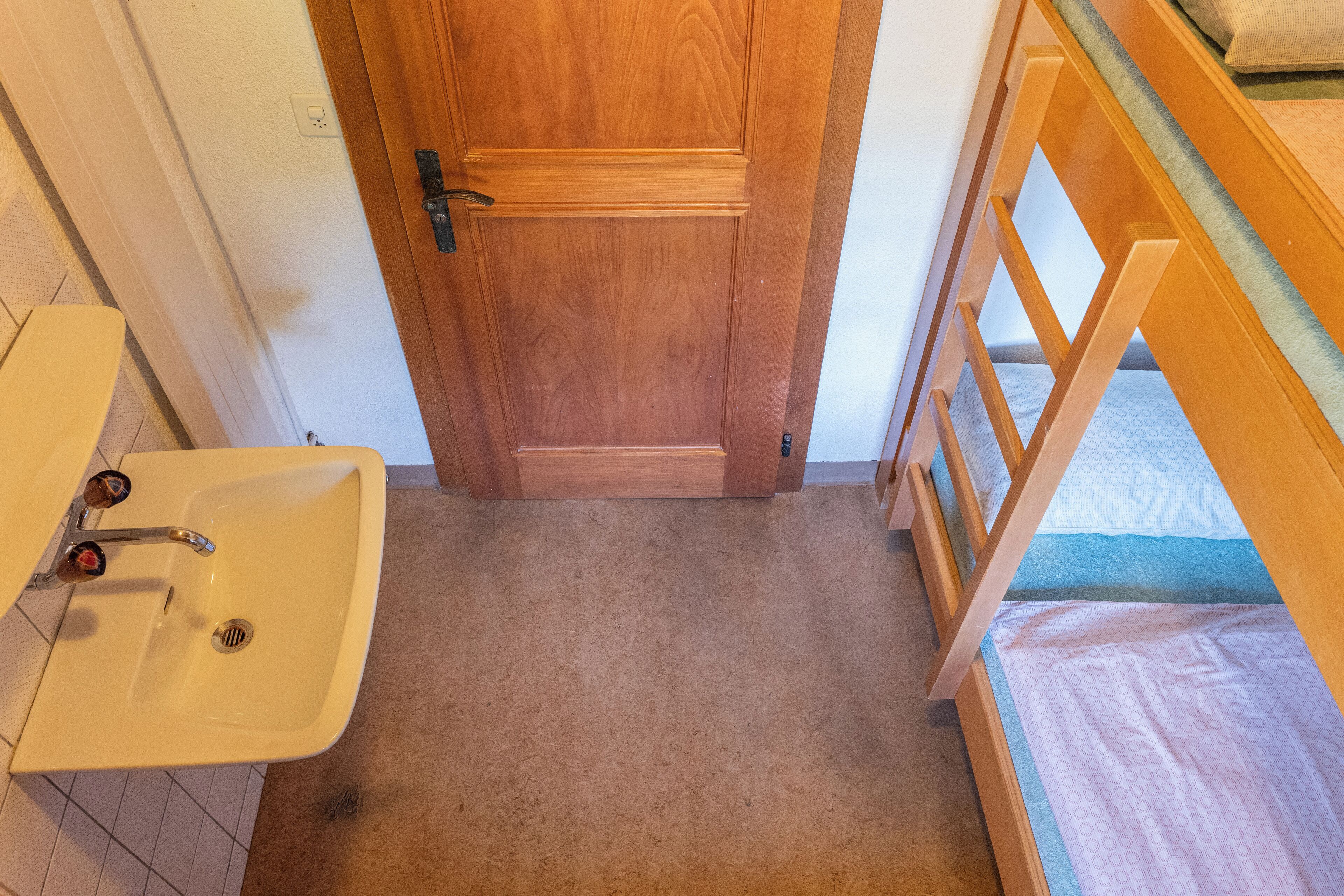 A small bathroom with a wooden door, a white sink, and a bunk bed with a mattress.