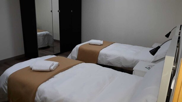A hotel room with two beds, each with white bedding and brown headboards. There is a mirror on the wall and a lamp on the side. - Europa House Village (San Vicente del Raspeig)