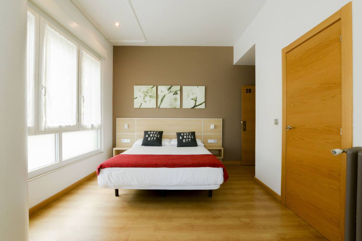 A bedroom with a bed, a wooden floor, a large window, and a door.