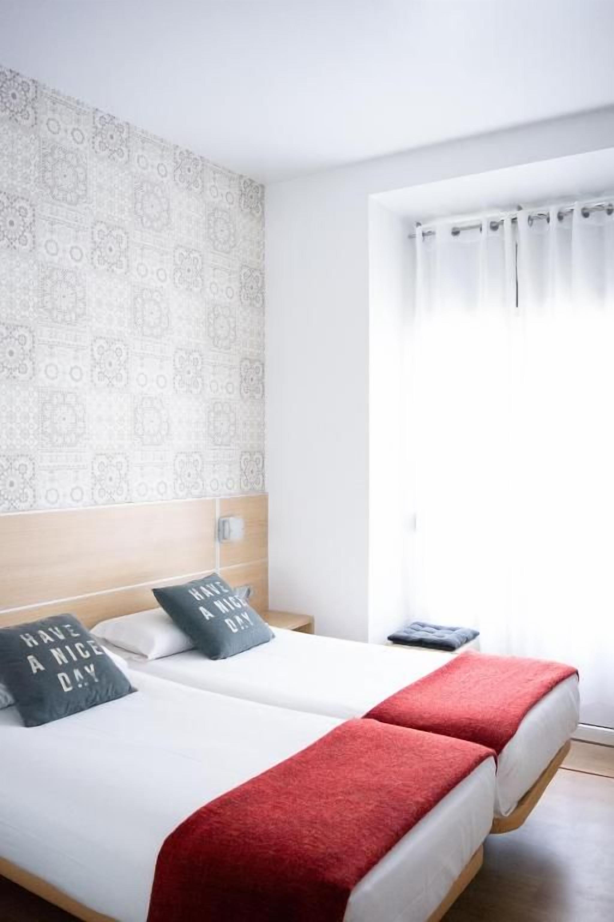 A hotel room with two beds, a red throw blanket, patterned wallpaper, and a window with curtains.