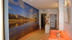 A hotel lobby with a large mural of a river and buildings, a reception desk, and an orange seating area. - Pensión Kursaal (San Sebastián)