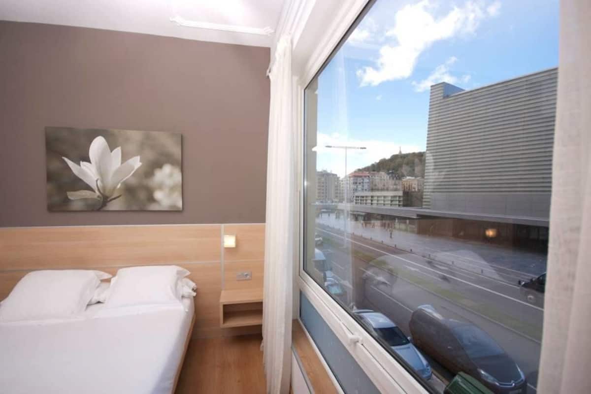 A hotel room with a bed, a bedside table, a nightstand, a window with a view of a cityscape and a road.