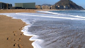 A beach with footprints in the sand, a building with a glass facade, and a hill with a statue in the background. - Pensión Kursaal (San Sebastián)
