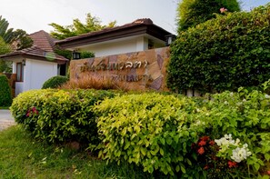 A sign for a hotel with a stone facade and lush greenery in front. - Baan Suan Nuanta (Samut Songkhram)