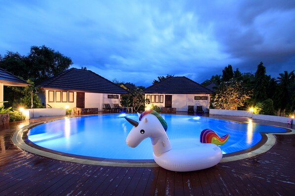 A swimming pool with a unicorn float, surrounded by wooden decking and two buildings with dark roofs. - Baan Suan Nuanta (Samut Songkhram)