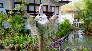 A welcome sign with a bird sculpture, surrounded by lush greenery and a small pond. - Baan Suan Nuanta (Samut Songkhram)