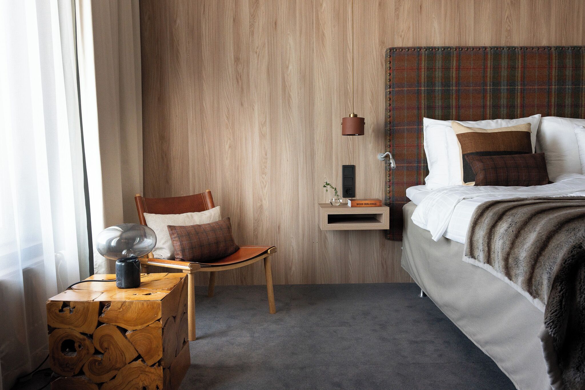 A hotel room with a wooden headboard, a bed with white linens, a wooden side table with a lamp, and a chair.