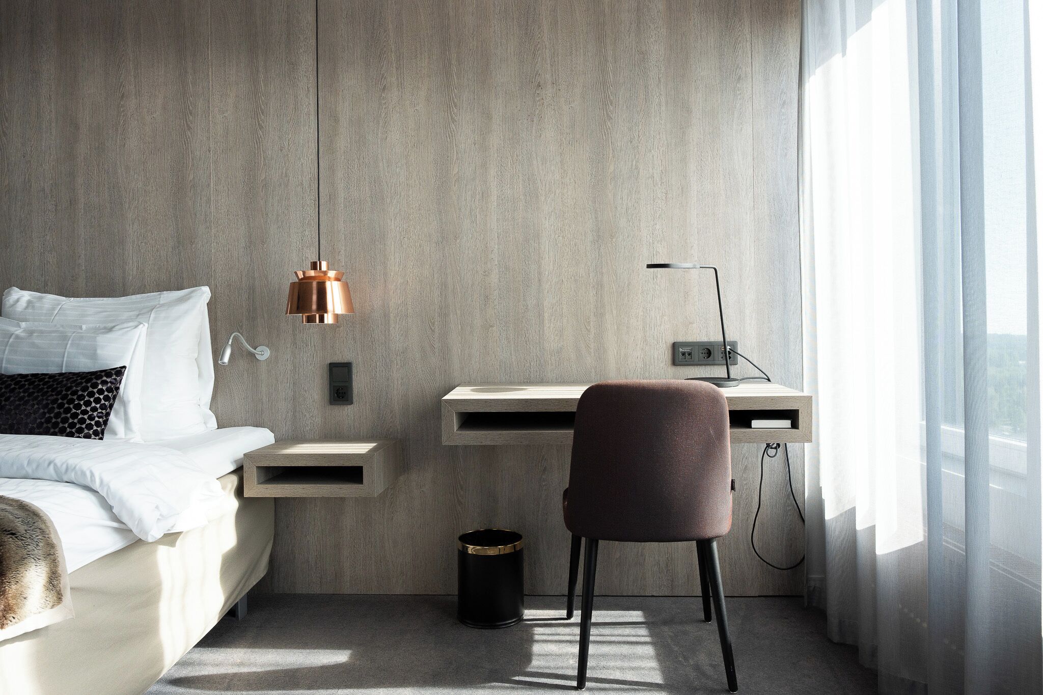 A modern hotel room with a bed, a desk, a chair, a lamp, and a window.