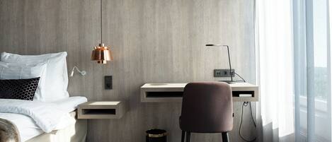 A modern hotel room with a bed, a desk, a chair, a lamp, and a window.