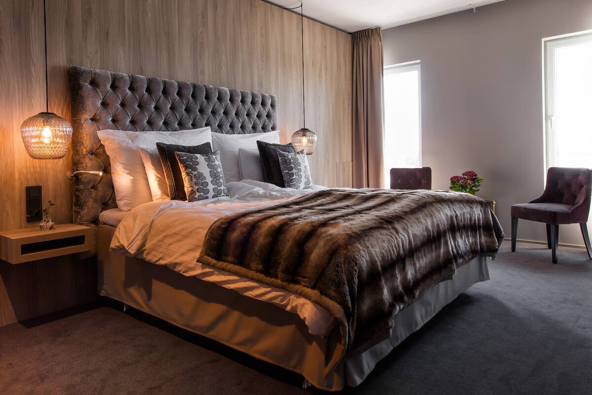 Een slaapkamer met een gestoffeerd hoofdeinde, een groot bed met meerdere kussens, een nachtkastje met een lamp, een stoel en een raam met gordijnen.