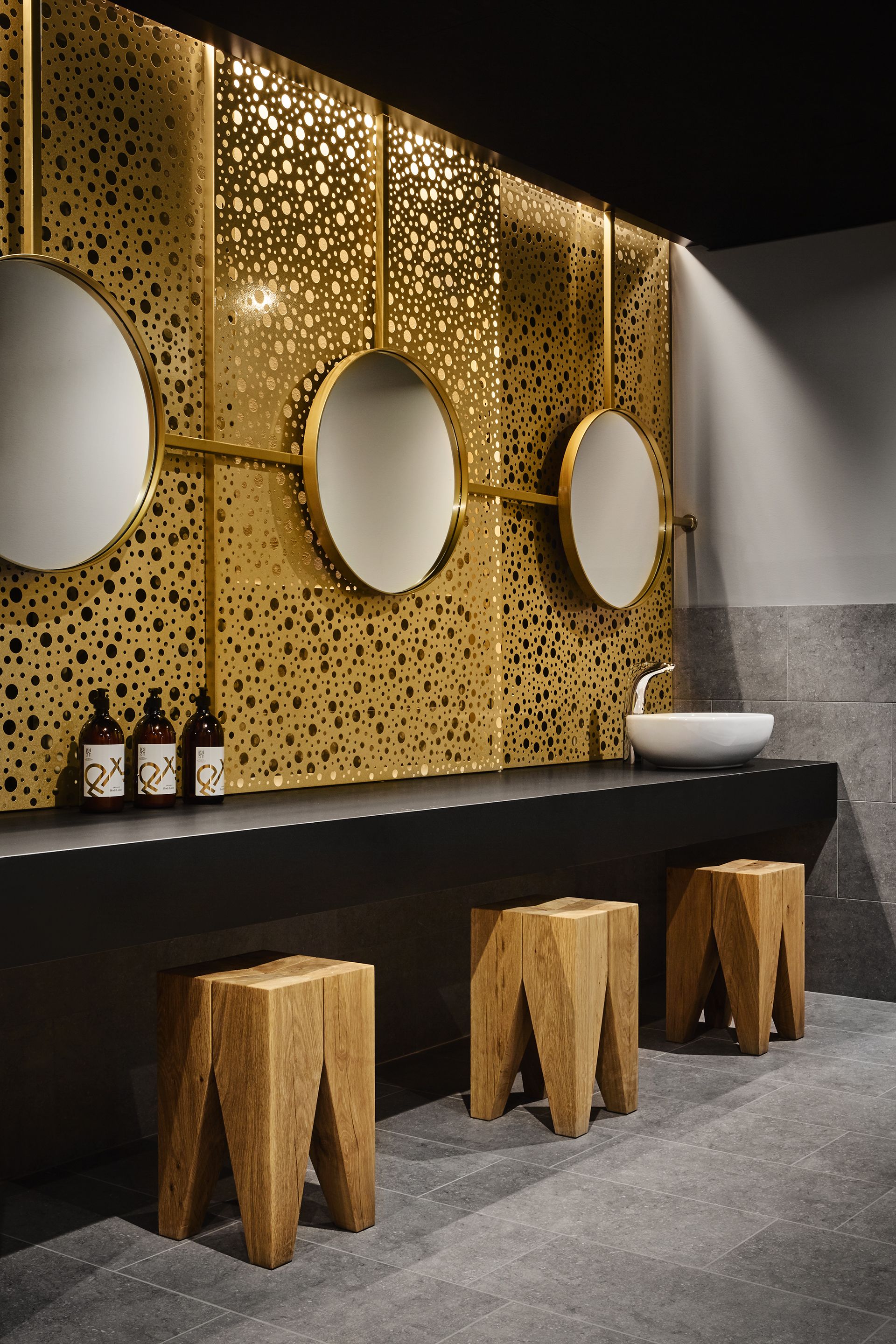 A modern bathroom with a perforated gold wall, three round mirrors, a white sink, and wooden stools.