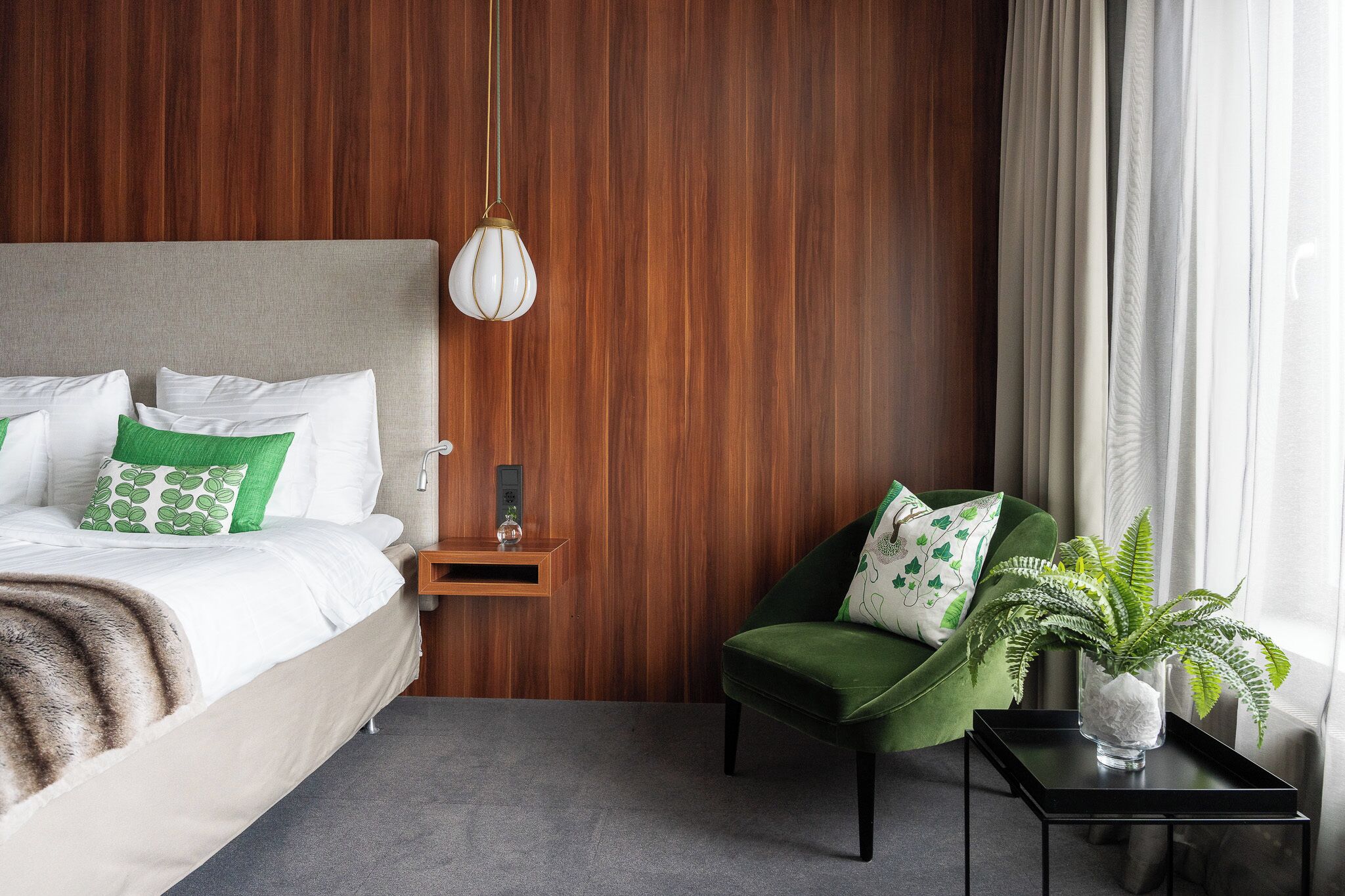 A hotel room with a wooden panel wall, a bed with white bedding and green pillows, a green armchair, a small table with a vase of green plants, and a hanging light fixture.