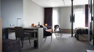 Quarto de hotel moderno com uma escrivaninha, duas cadeiras, um sofá, uma cama, uma mesinha de cabeceira com abajur e uma televisão.
