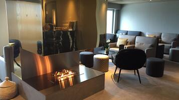 A modern lounge area with a central fire feature, comfortable seating, and a large mirror reflecting the space.