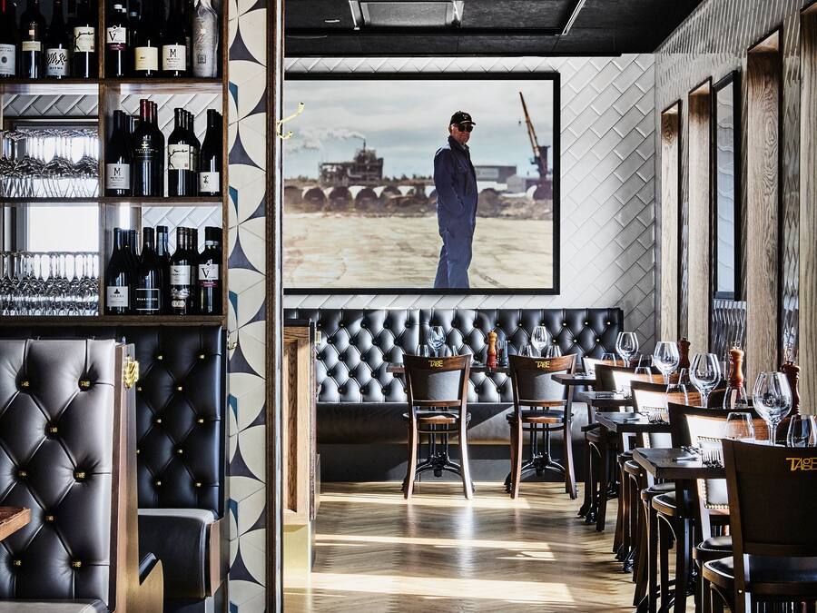 Zona de bar moderna con suelo de madera, asientos de cuero y una pantalla grande que muestra a una persona.
