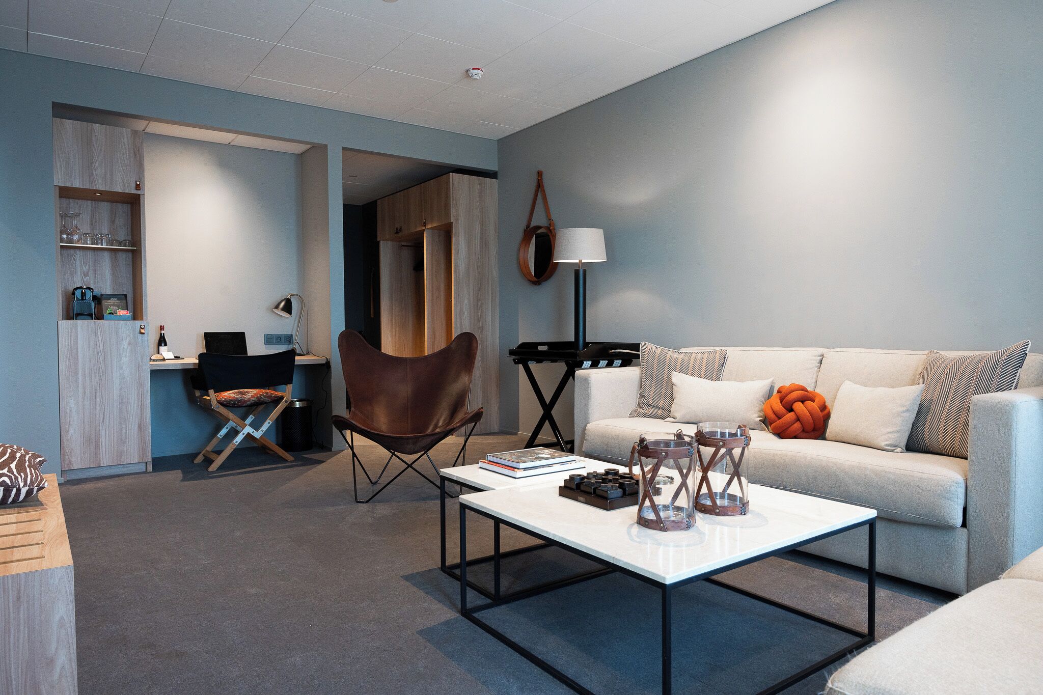 A modern living room with a sofa, coffee table, and a chair. There is a desk with a computer and a lamp. The room has a light blue wall and a wooden cabinet.