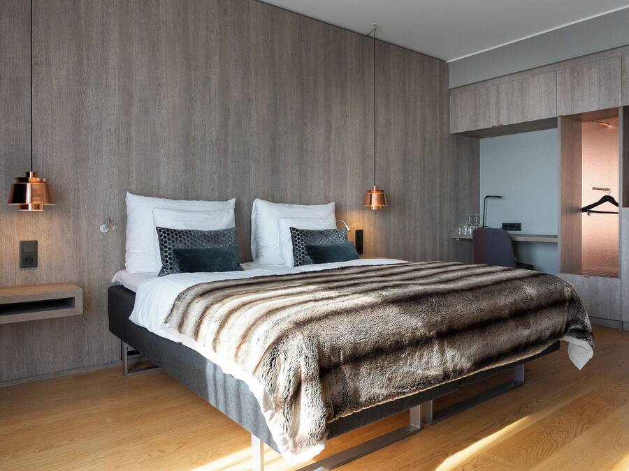 A modern bedroom with a wooden wall, a bed with a striped blanket, bedside lamps, and a built-in wardrobe.