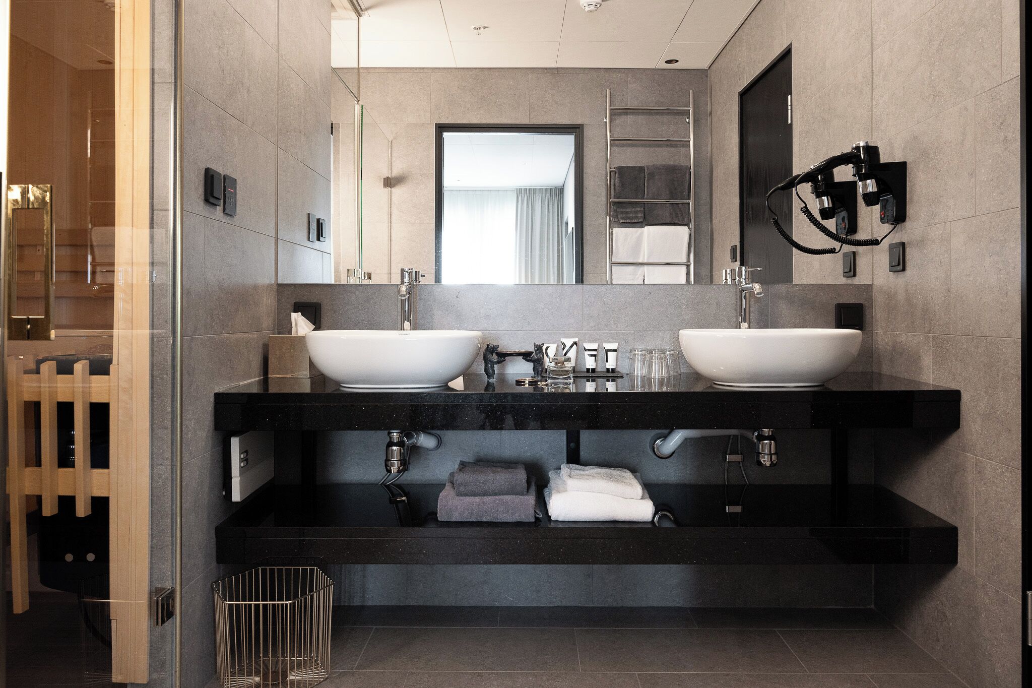 A modern bathroom with two sinks, a mirror, towel racks, and a trash bin.