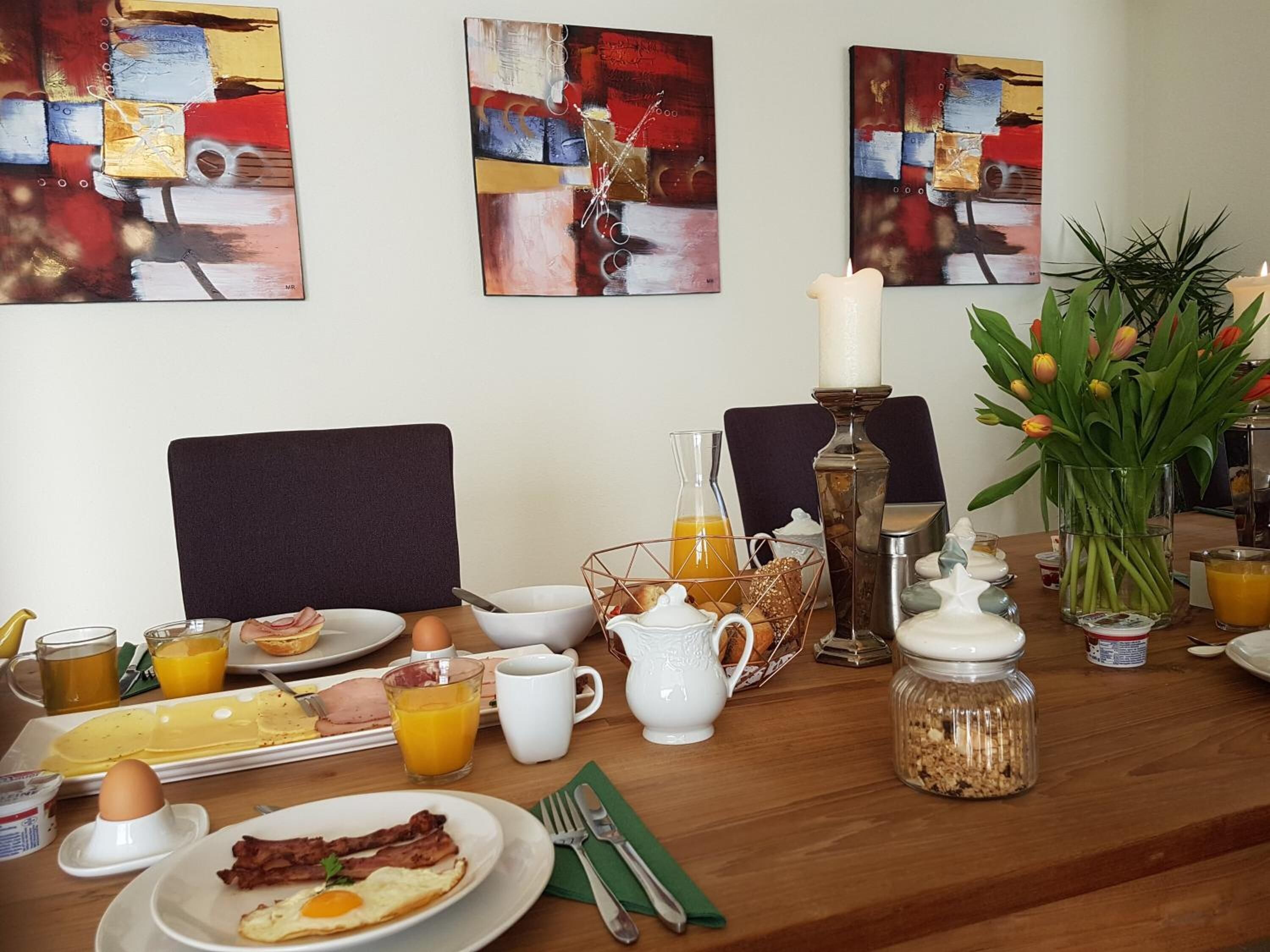 Ein Frühstückstisch mit Tellern mit Eiern, Speck und Toast, einer Tulpenvase und abstrakten Gemälden an der Wand.