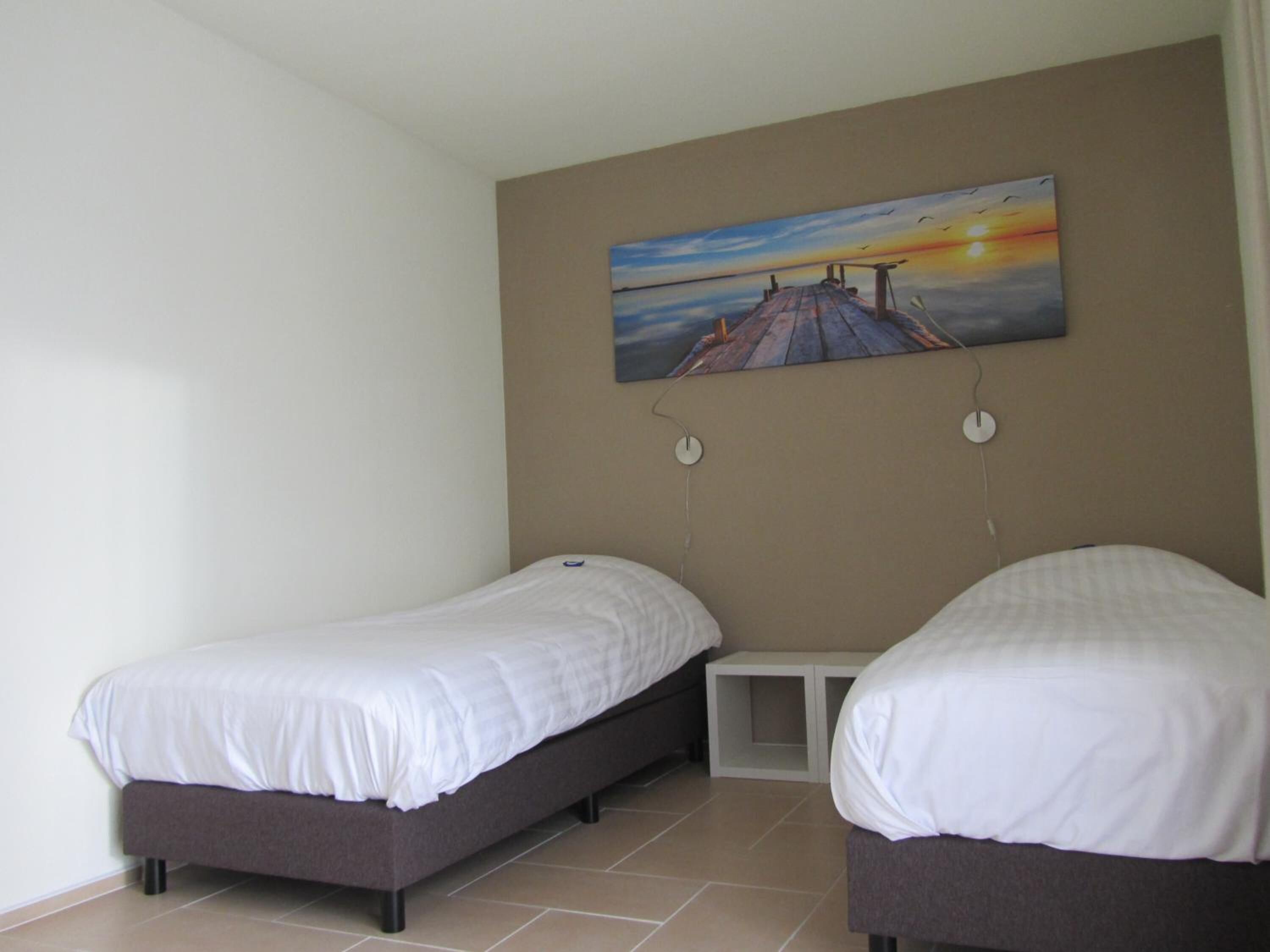 Een slaapkamer met twee eenpersoonsbedden, een klein nachtkastje en een schilderij van een ondergaande zon aan de muur.