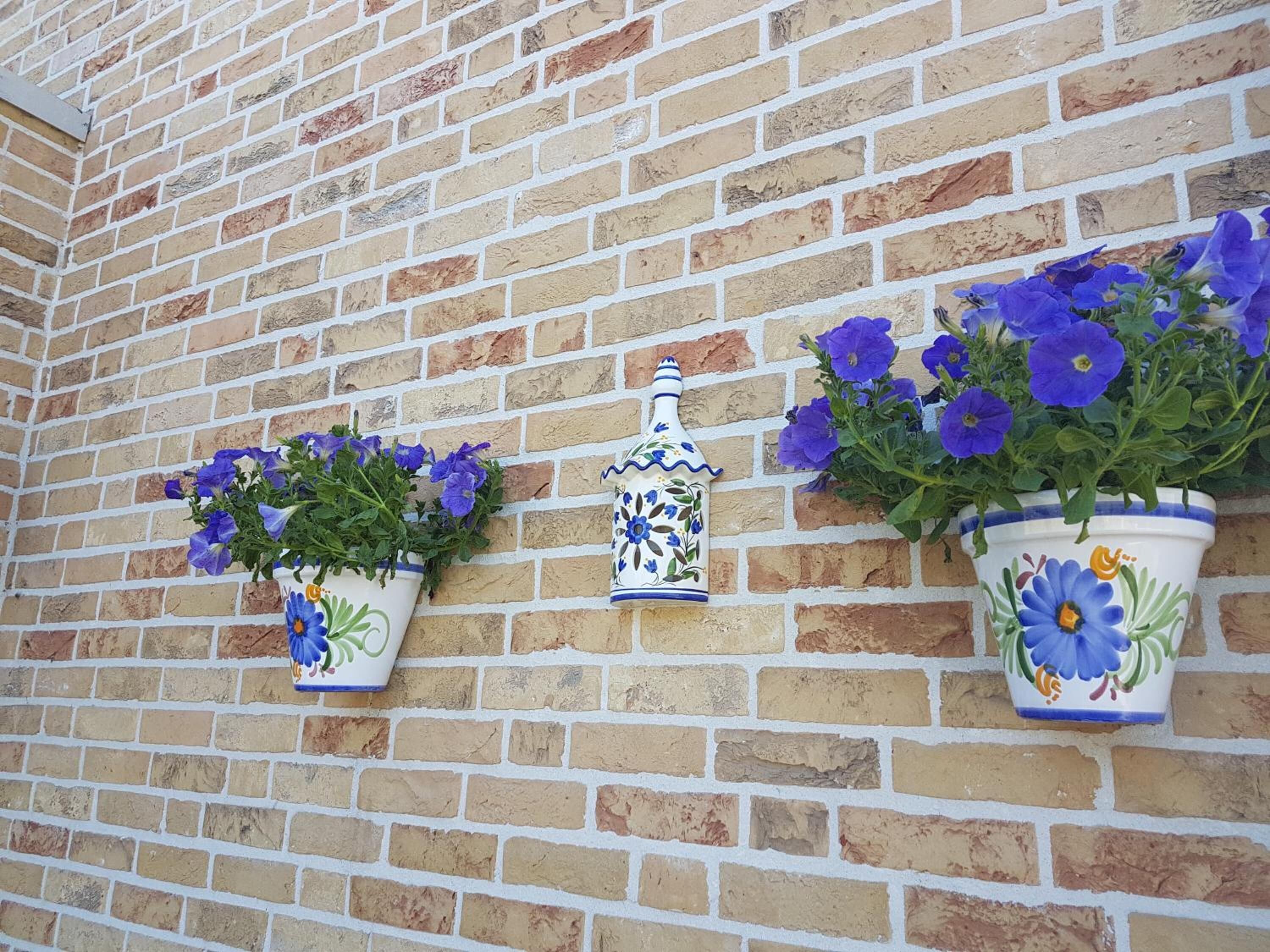 En mur af mursten med tre blomsterpotter, hver med lilla blomster, samt et dekorativt keramikstykke.