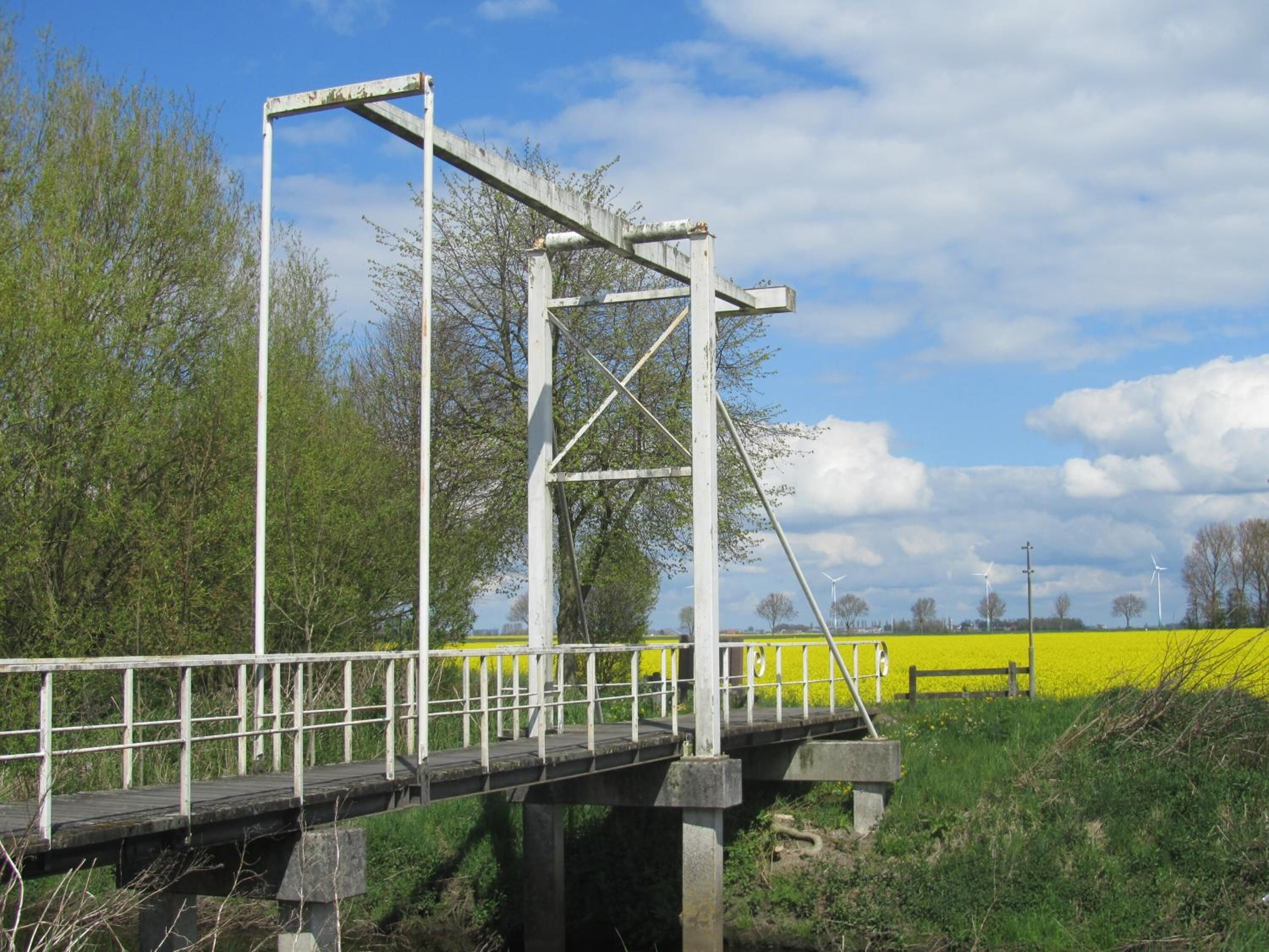 Een metalen brug met een houten looppad, omgeven door groen en velden.