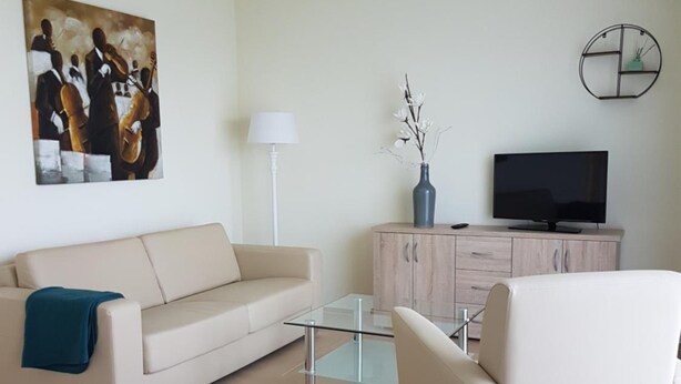 Un salón moderno con un sofá color beige, una mesa de centro de cristal, un sillón blanco, un televisor sobre un mueble de madera y un cuadro en la pared.