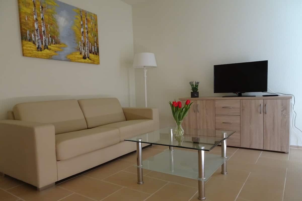 A living room with a beige sofa, a glass coffee table, a painting on the wall, a TV on a wooden cabinet, and a vase with red flowers.