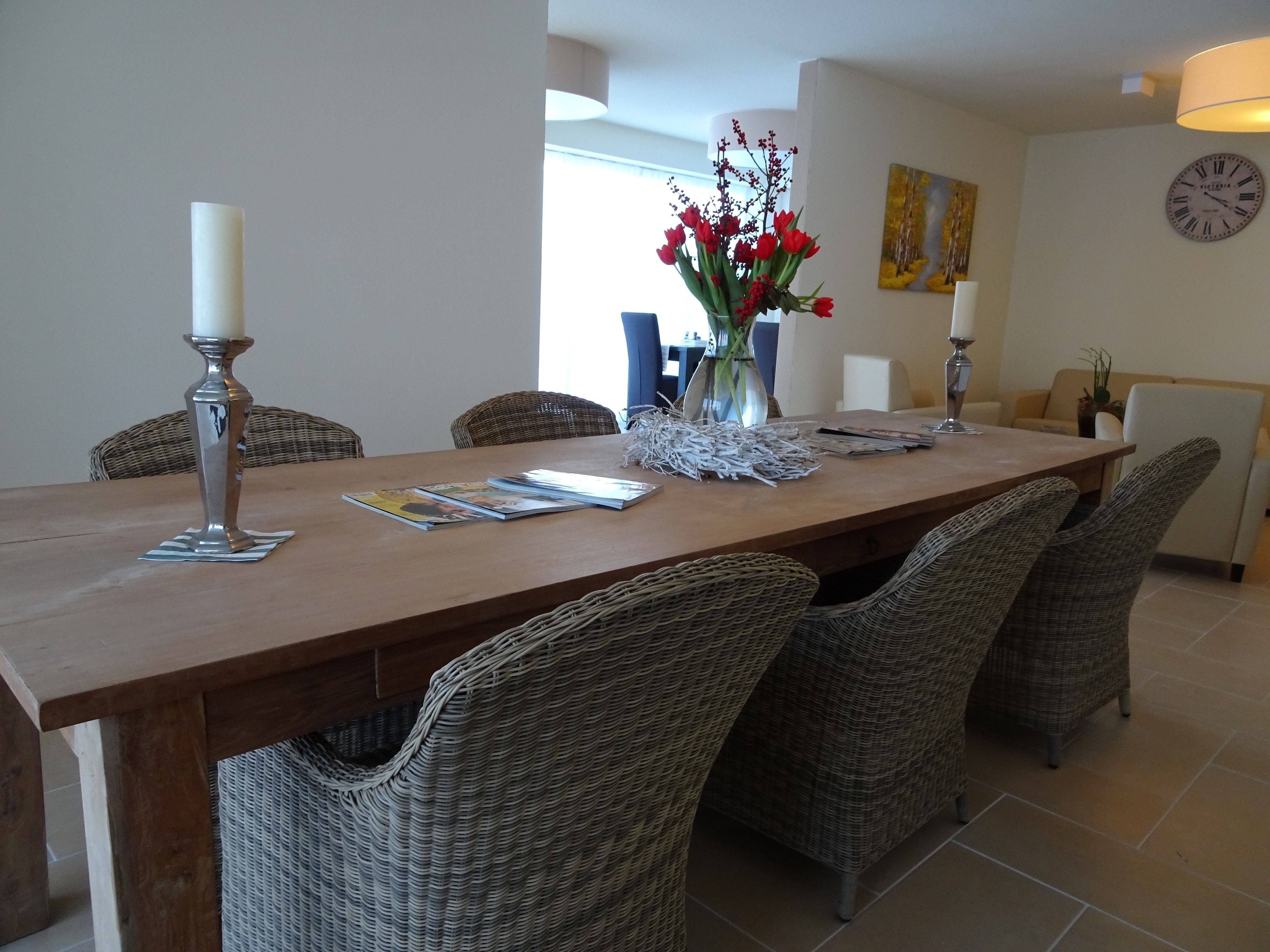 A dining area with a wooden table, wicker chairs, a vase with red flowers, and a clock on the wall.