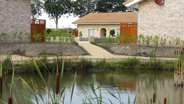 Un edificio in mattoni con tetto in tegole, un vialetto lastricato che conduce all'ingresso e uno stagno con giunchi in primo piano.