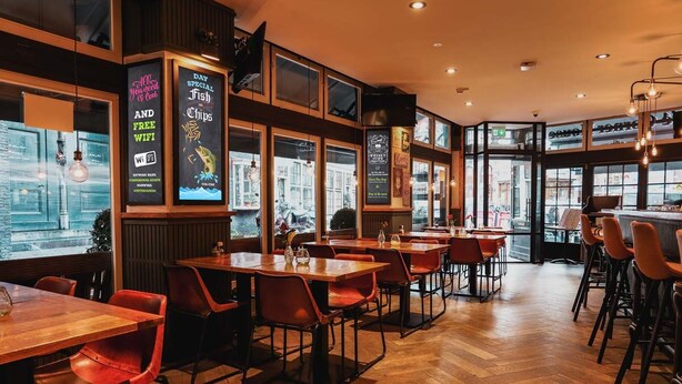 A restaurant interior with wooden tables and red chairs, large windows, and a herring sign. - Hotel Corner House (Amsterdam)