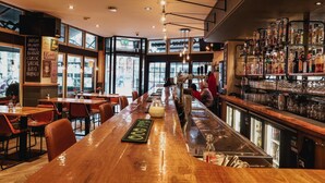 A bar area with a long wooden counter, seating on both sides, and a large window providing a view of the street. - Hotel Corner House (Amsterdam)
