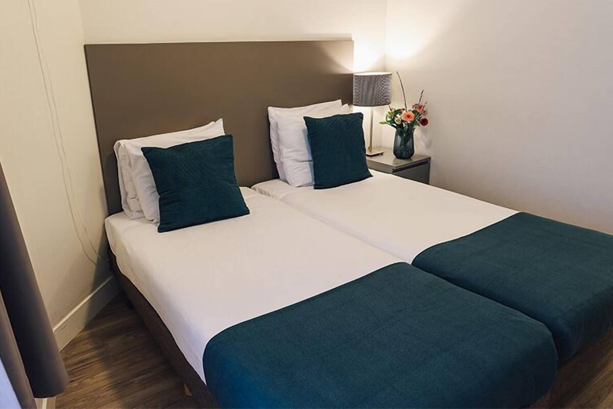 A double bed with two pillows, a bedside table with a lamp and flowers, and a wooden floor.