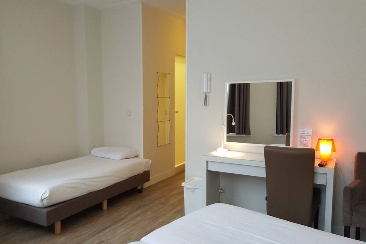 A small, clean room with a single bed, a desk with a chair, a mirror, and a bedside lamp.