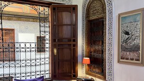 A room with a purple sofa, a small table, and a lamp. There is a window with a decorative iron grille and a door with intricate woodwork. - Dar Attajalli (Fes)