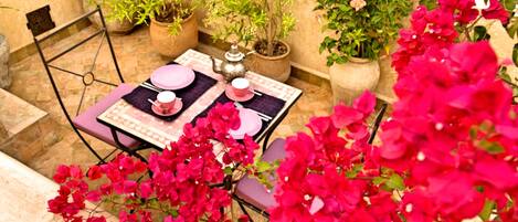Una terraza con una mesa para dos, rodeada de macetas con plantas y coloridas flores de buganvilla.